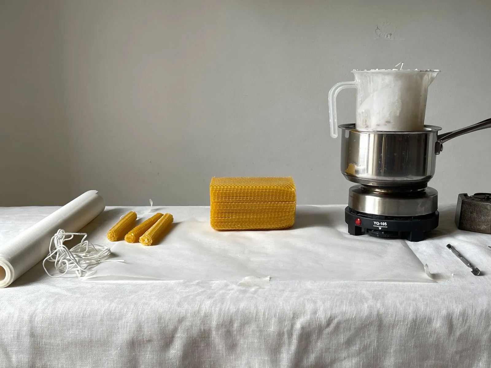 A block of raw beeswax, a coil of cotton wick and a pot of melted wax on a linen cloth